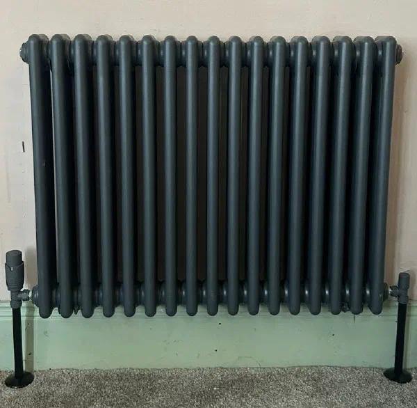 Anthracite grey column radiator wall-mounted above a painted skirting board, with matching dark valves and floor-standing supports