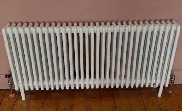 White three-column radiator fitted against a pink wall on stripped pine floorboards, with traditional valves and copper tails
